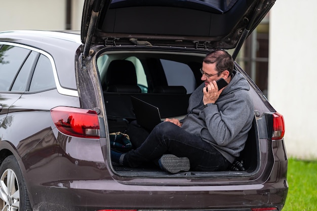 Photo a man sitting in the open trunk of a car and working on a laptop technology to work remotely