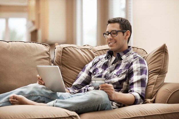 A man sitting on a sofa using a digital tablet and holding a credit card