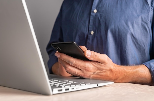 Man sitting at table with laptop and holding phone Businessman using smartphone for work or leisure in office at home Male texting message or surfing Internet