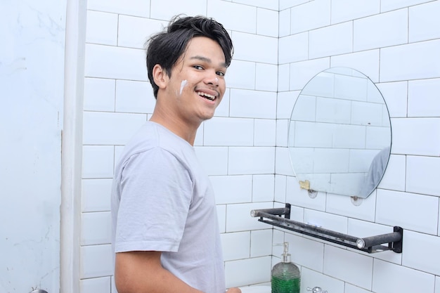 Man skincare concept Friendly attractive young man applying moisturising cream on face while standi