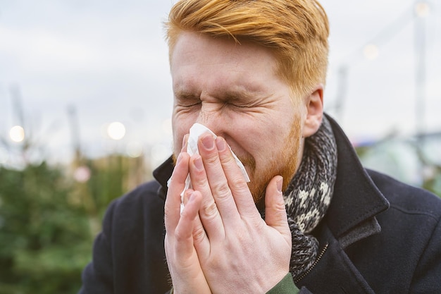 man sneezes and blows his nose in a handkerchief at winter time with allergy or an infection a virus. Portrait of redhead man outdoor.