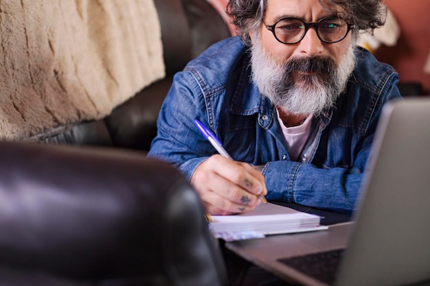 Man on a sofa with a laptop at home Hipster make note while studying online watching webinar podcast on laptop listening educational courseelearning learning concept