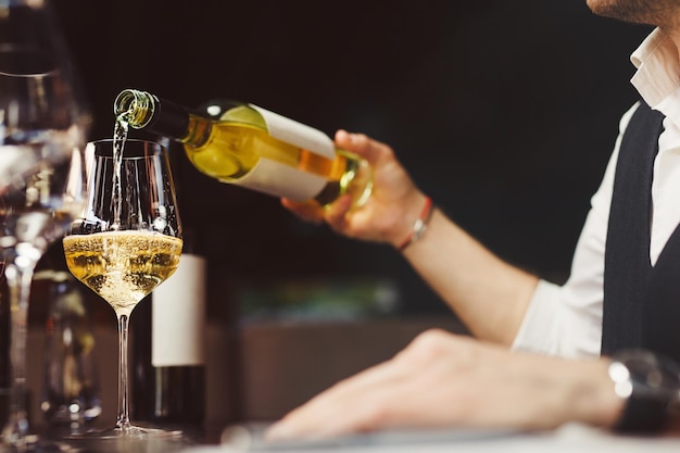 Man sommelier pours white wine into stylish wineglass sitting at table in restaurant close side view