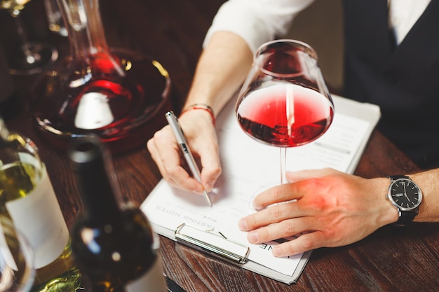 Man sommelier writes on clipboard with paper describing delicious red wine taste quality at table in luxury restaurant closeup