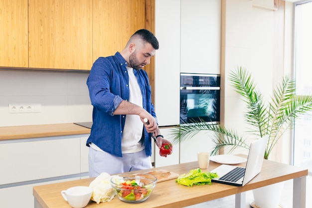 man spends a day at home prepares a breakfast of vegetables in the kitchen a man in home clothes and with a beard uses a laptop to learn online cooking