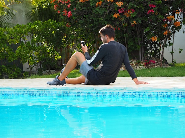 A man in sportswear, sitting near the swimming pool in the hotel, uses a cell phone