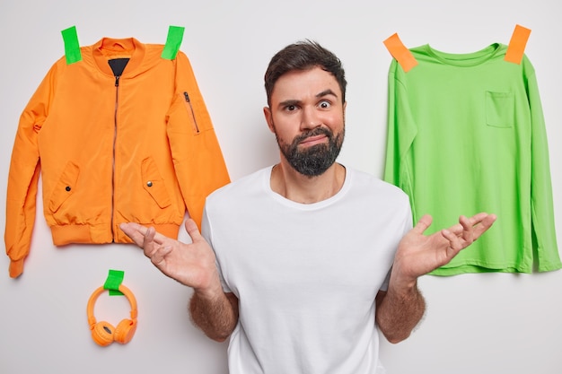  man spreads palms feels hesitant about what to wear thinks about new purchase wears casual t shirt poses on white with plastered clothes and accessories