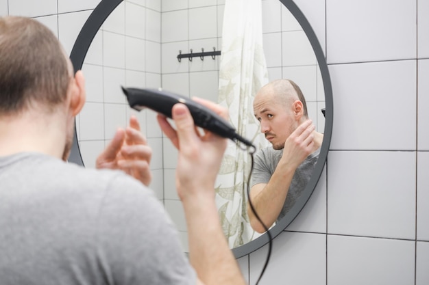 A man standing against mirror in the bathroom holding a trimmer and cutting hair by himself