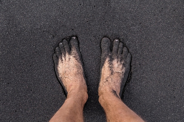 A man standing barefoot on volcanic black sand on the Tenerife beach. Two feet covered by the sand.