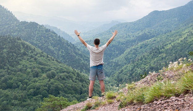 Man standing on the slope of the mountain raised his hands up and admires the beautiful view of the mountain panorama