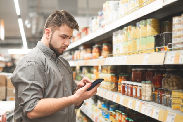 Man stands in the aisle of the supermarket at shelves with canned vegetables and uses a smartphone