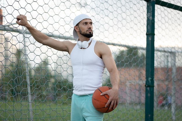 A man stands near a fence on the street, holding a basketball in his hand