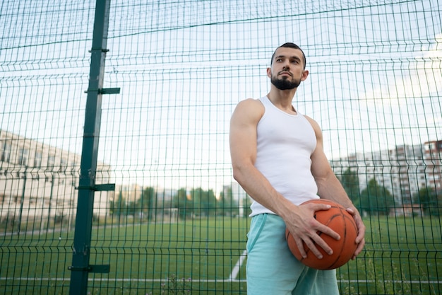 A man stands near a fence on the street, holding a basketball in his hand