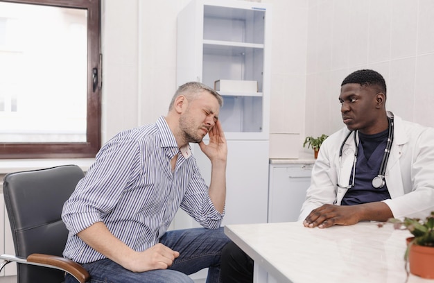 A man suffering from a severe headache is sitting at an AfricanAmerican doctor's appointment