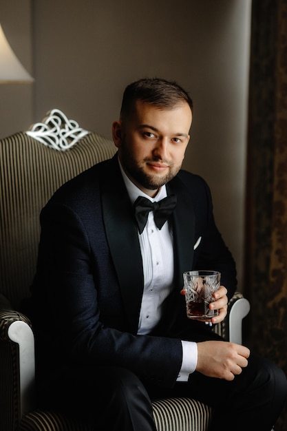 A man in a suit sits in a chair with a glass of whiskey.