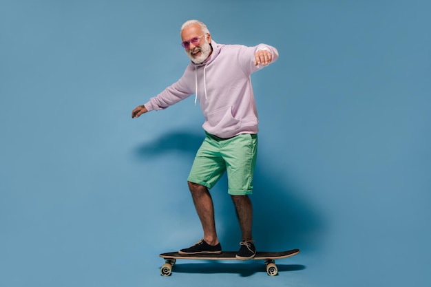 Man in sunglasses hoodie and shorts riding skateboard Positive adult guy with white hair in lilac sweatshirt and green shirt posing