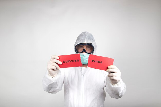 Man in surgical mask wearing white protective suit and a red sign with the word coronavirus split in half isolated in studio on white background. Prevention against coronavirus.