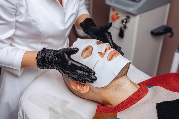 A man takes the procedure from a cosmetologist who makes him a tissue mask with collagen and vitamins