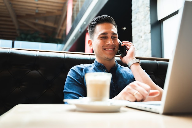 Man talking at the mobile phone while using computer laptop sitting at cafe table