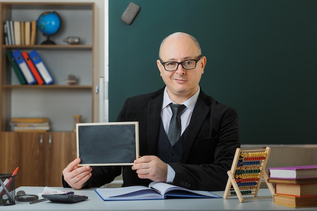 Man teacher wearing glasses sitting at school desk in front of blackboard in classroom showing chalkboard explaining lesson explaining lesson with smile on face