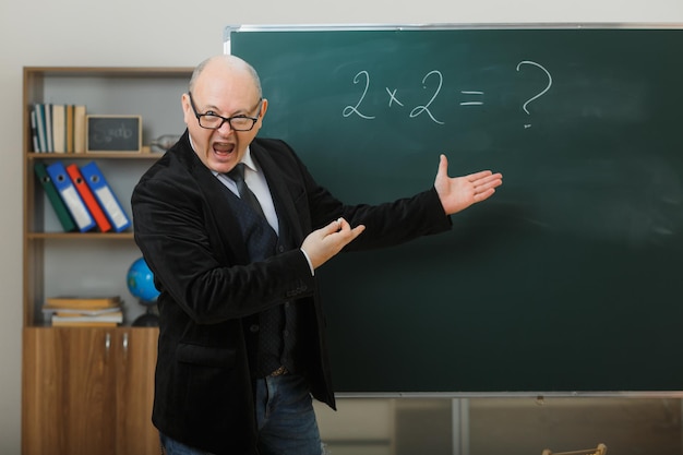 Man teacher wearing glasses standing near blackboard in classroom explaining lesson with aggressive expression being angry and mad