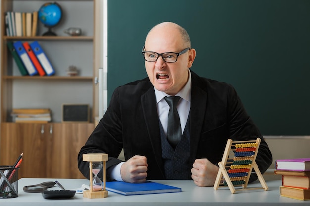 Man teacher wearing glasses with class register sitting at school desk in front of blackboard in classroom using abacus angry and frustrated clenching fists shouting