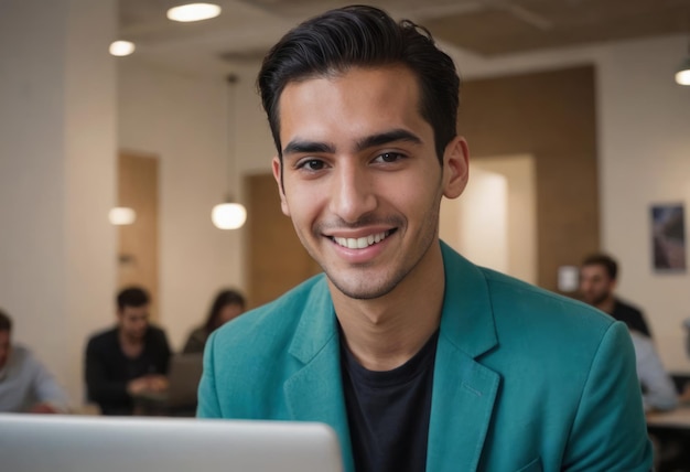 A man in a teal blazer smiles gently while using a laptop in a busy office backdrop