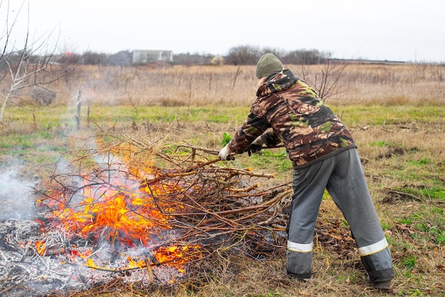 A man throws dry branches into a burning fire in a field Burning trash and branches in the garden