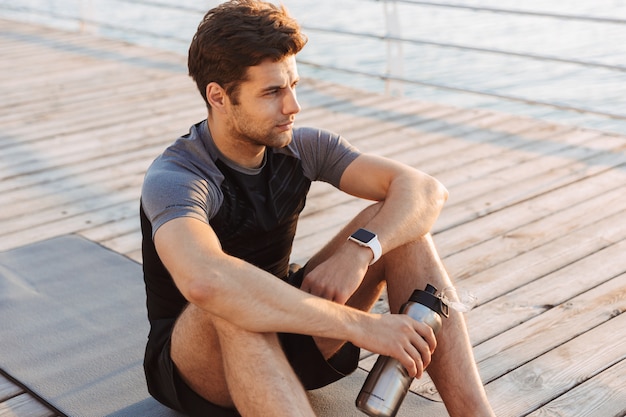 Man in tracksuit sitting on mat with thermos mug after workout on wooden pier, at seaside in morning