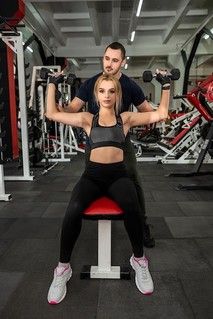 Man trainer helps her slim woman client exercising with heavy dumbbells at the gym Fitness and gym for health concept