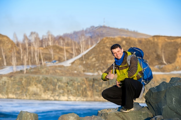 Photo man traveler with a backpack shows thumb on the lake