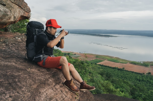 Man traveler with backpack using camera taking a photo on edge of cliff, on a top of the rock mountain