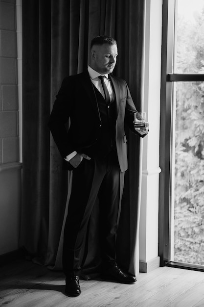 a man in a tuxedo stands in front of a window with a glass of wine