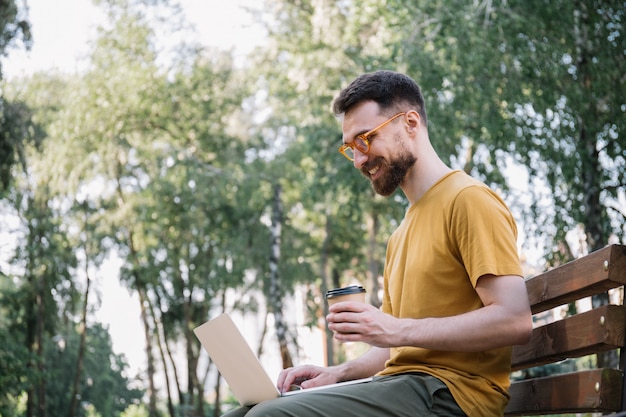 Man using laptop, holding cup of coffee, sitting on bench. Freelancer copywriter working in park