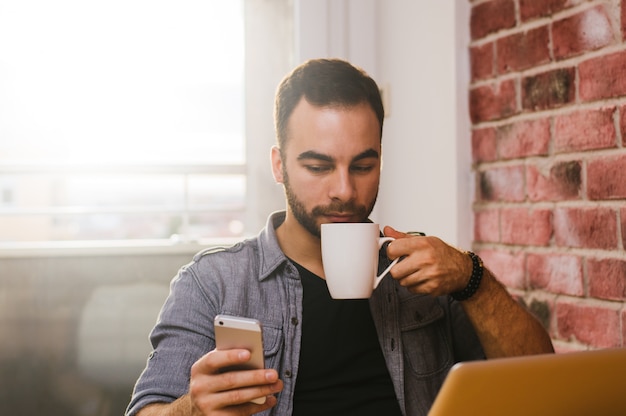 Man using mobile and laptop at home in the morning, drinking coffee