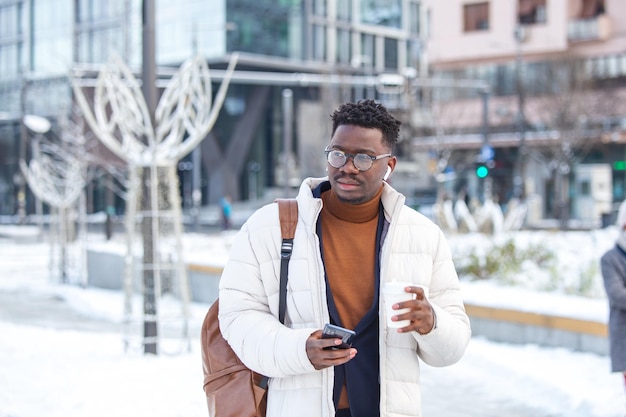 Man using phone with wireless headphones walking in the city and drinking coffee