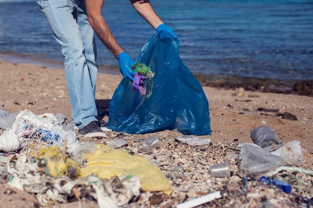 Man volunteer in blue t-shirt with big bag for trash collecting garbage on beach. Environmental pollution concept