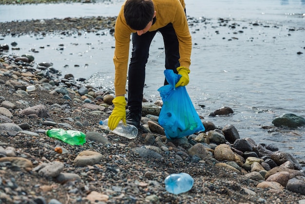 Man volunteer picking up plastic trash on stone beach Male collecting used plastic bottles in trash bag Earth Day