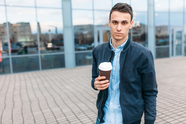 A man walks around the city and drinking coffee from a paper Cup, a handsome guy walks around and resting, the guy on the background of a modern building, office