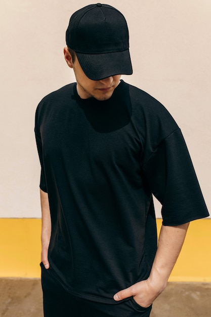 Man wearing black tshirt and a black baseball cap with on an outdoors wall background