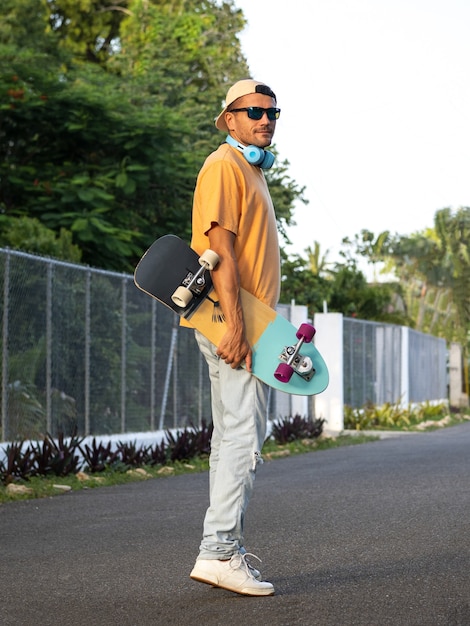 Man wearing cap, sunglasses and headphones, walking with skateboard.