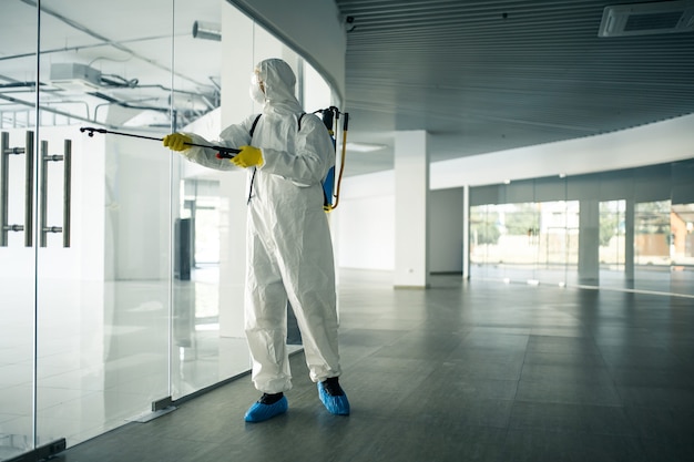 A man wearing disinfection suit spraying with sanitizer the glass doors' handles in an empty shopping mall to prevent covid-19 spread. Health awareness, clean, defence concept.