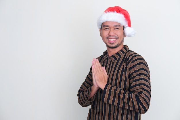 A man wearing Javanese costume and christmas hat smiling with hand praying pose