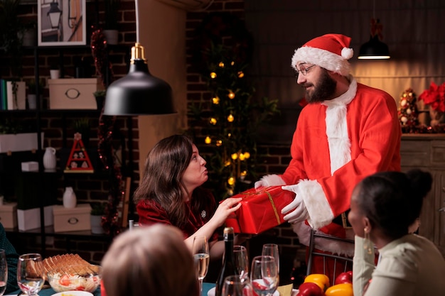 Man wearing santa claus costume giving woman christmas gift, happy wife getting present giftbox from husband. Xmas night celebration with big family at festive dinner table, winter holidays greeting