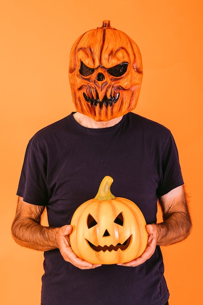 Man wearing scary pumpkin latex mask with blue t-shirt, holding 'Jack-o-lantern' pumpkin on orange background. Halloween and days of the dead concept.