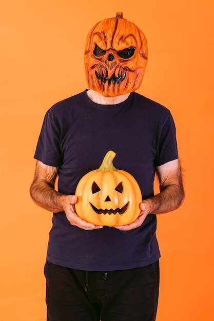 Man wearing scary pumpkin latex mask with blue t-shirt, holding 'Jack-o-lantern' pumpkin on orange background. Halloween and days of the dead concept.