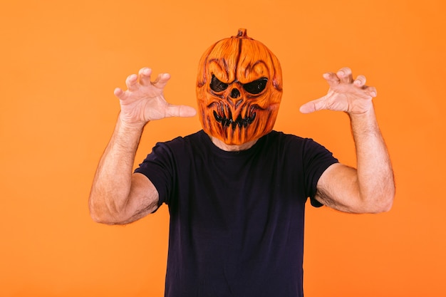 Photo man wearing scary pumpkin latex mask with blue t-shirt scares with his hands, on orange background. halloween and days of the dead concept.