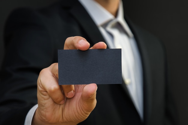 Man wearing a suit holding white business card on black wall surface.