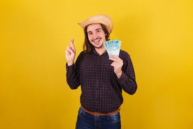 Man wearing typical clothes for party Junina Holding money bills Brazilian real and fingers crossed wishing and cheering finance