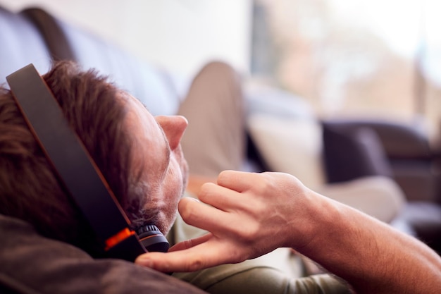 Man Wearing Wireless Headphones Listening To Music Lying On Sofa At Home Wearing Pyjamas